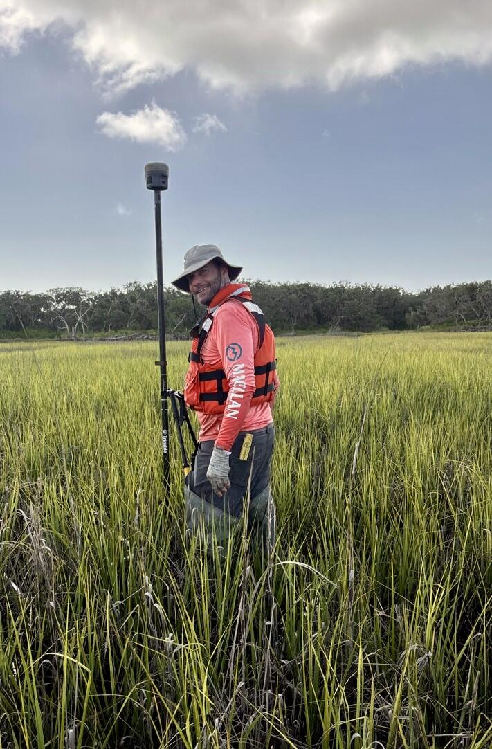 USGS scientist, Nicholas Enwright, holds survey equipment on a tall pole as he stands in a grassy marsh. He is wearing a brown bucket hat, a red long-sleeved shirt, and a orange life jacket. Trees can be seen in the distance under a blue sky with scattered clouds.