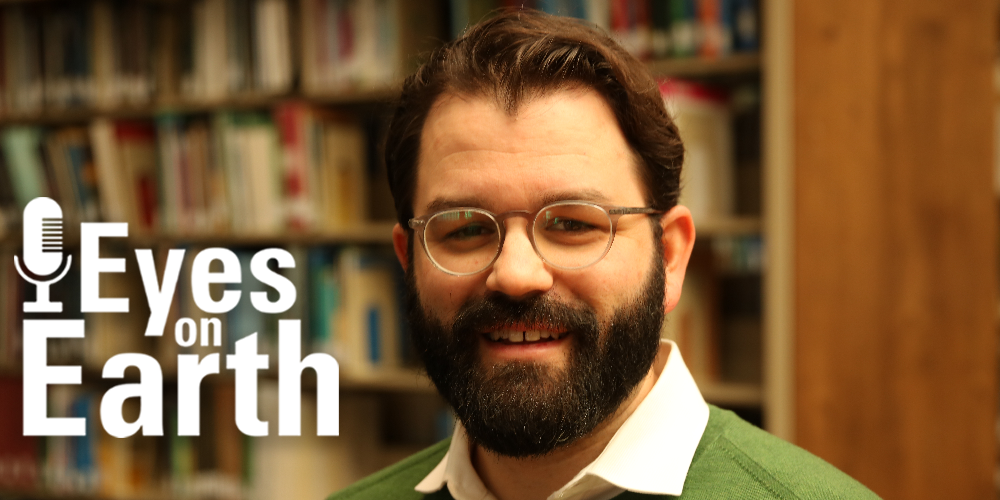Mugshot of bearded man with bookshelves behind him and a little white text