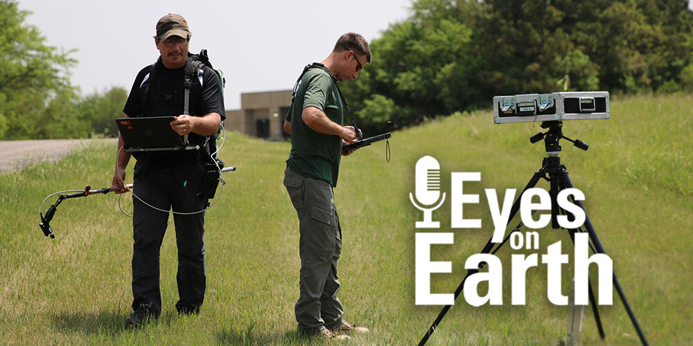 Two men are working with equipment in a grassy area with trees and a building in the background and a road to the left