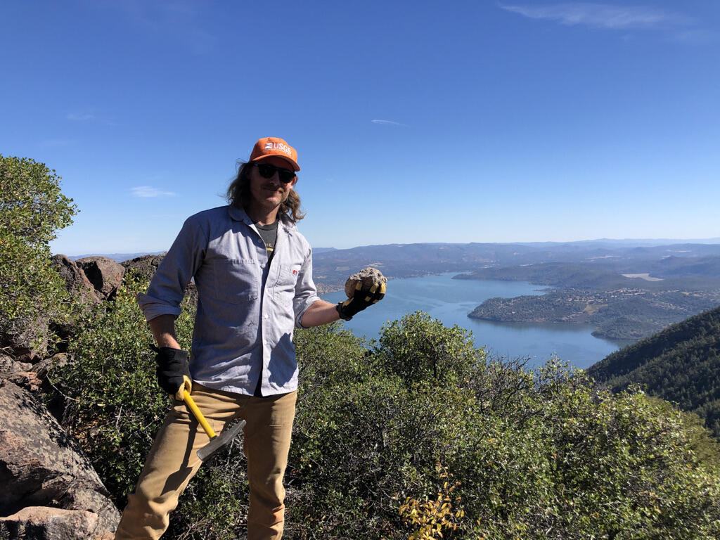 A male geologist in an orange ballcap and blue button-down shirt holds a rock hammer in one hand and a rock in the other.