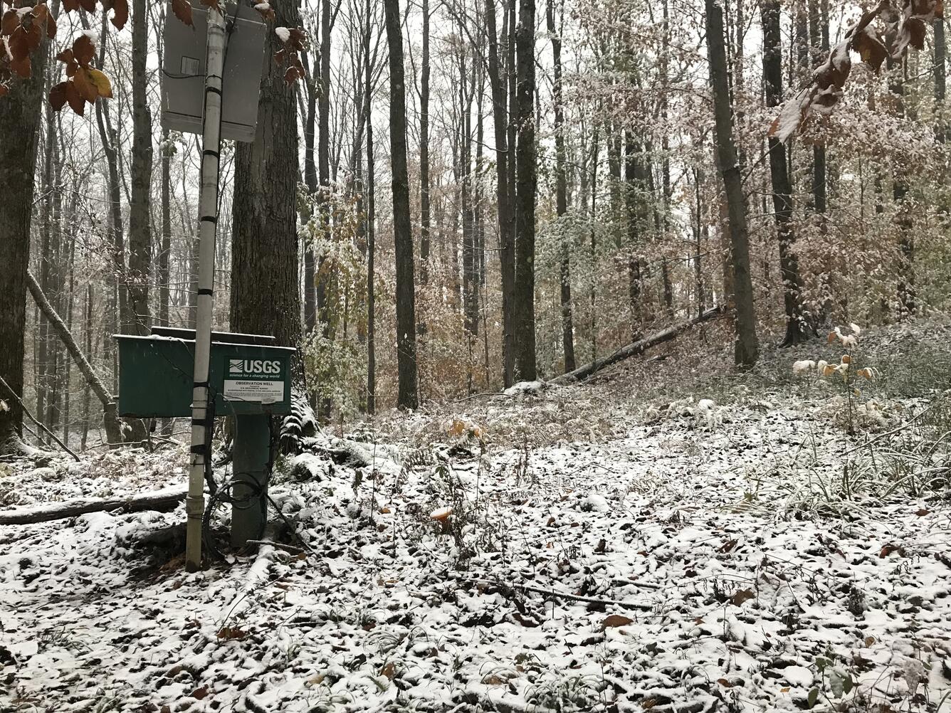Photograph showing a groundwater monitoring well in a winter snow scene on a forested hillside..