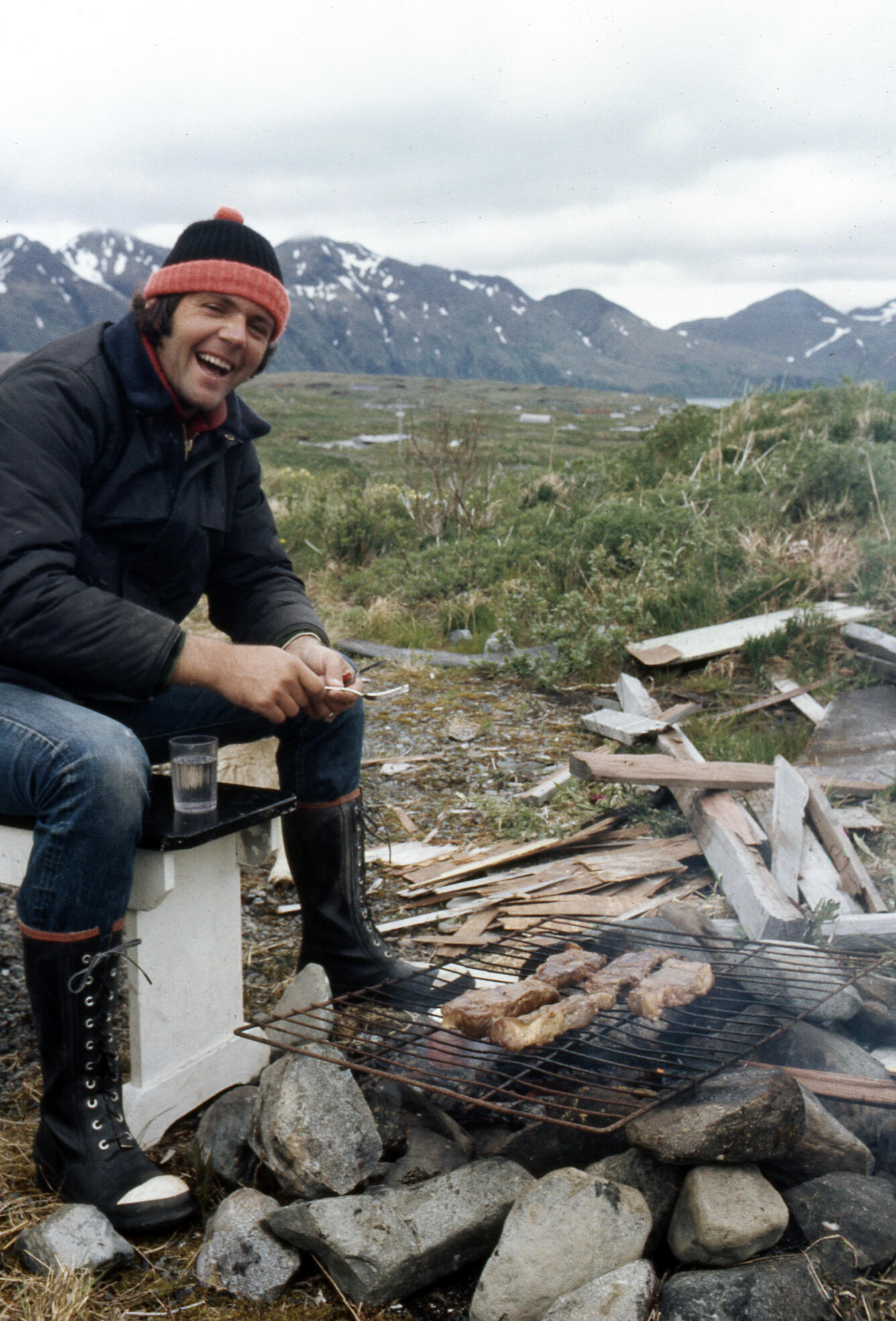 A young Jim Estes sits near the campfire with snowy mountains in the back, circa 1972