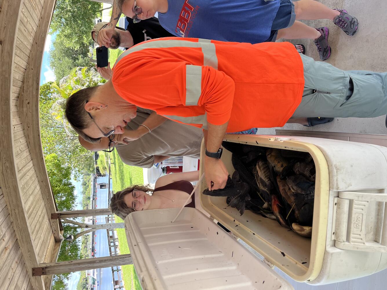A group of people stand around a cooler filled with nonnative fish as a man in an orange shirt gives identification guidance