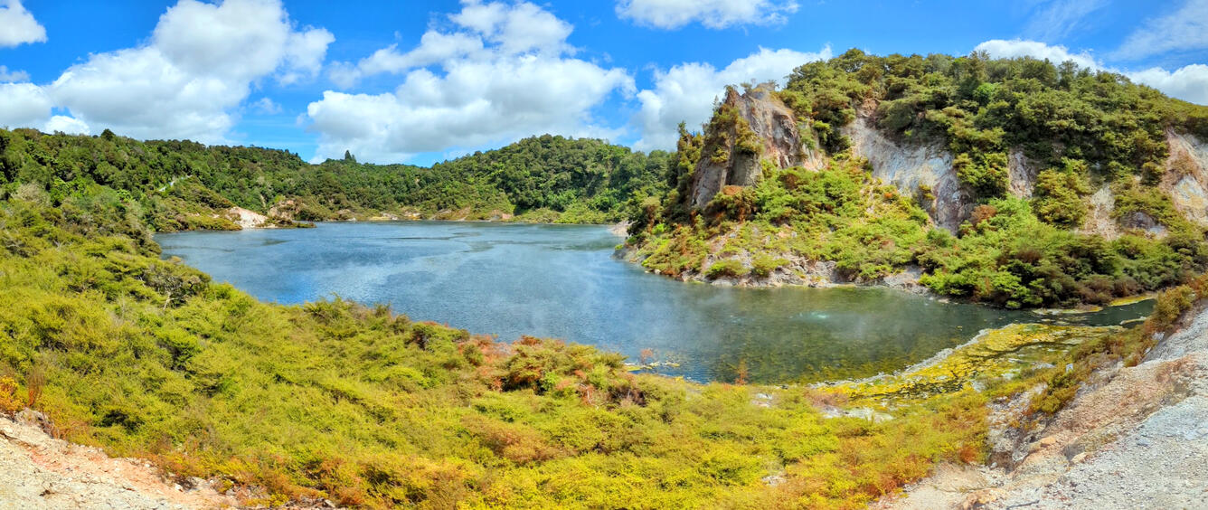 A small lake surrounded by scrubby green vegetation under a partly cloudy sky. The right side of the lake shore includes a tall mass of partly vegetated rock.