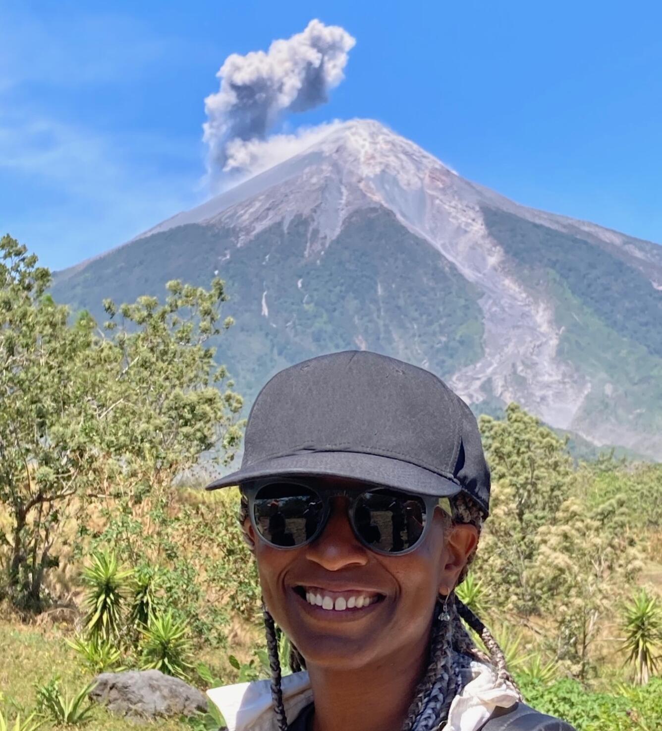woman with black baseball hat with a volcano erupting ash in the far background.