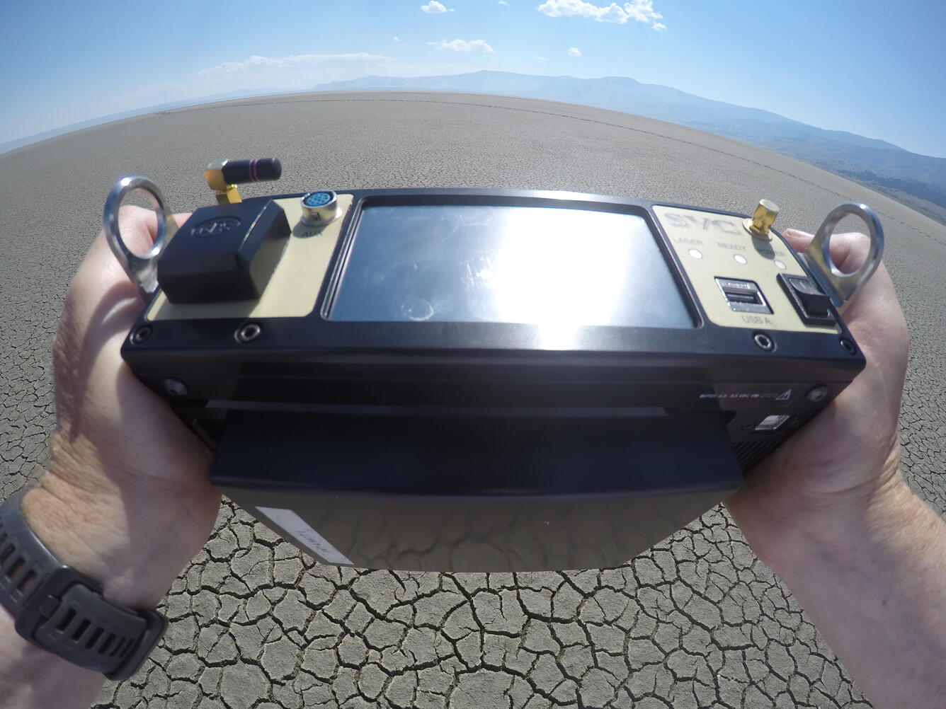 Person holds a rectangular device in both hands pointing it at the dry desert landscape.