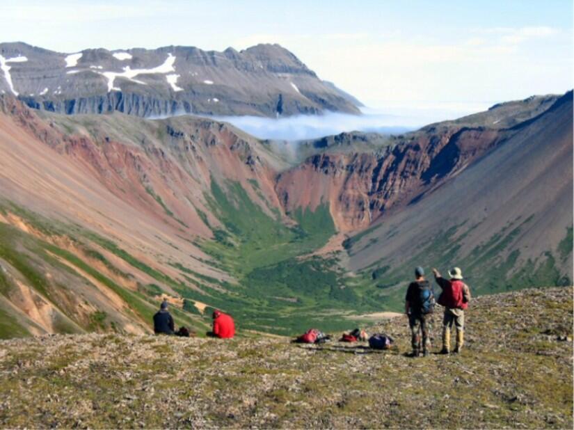 Two people standing by a ledge facing and gesturing away towards mountains in the distance; packs and two people on ground