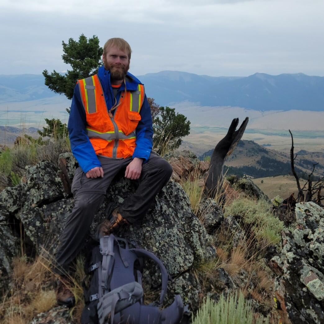 Geologist sitting on lichen covered bolder in orange safety vest and backpack by his feet. Tree and mountains behind him.