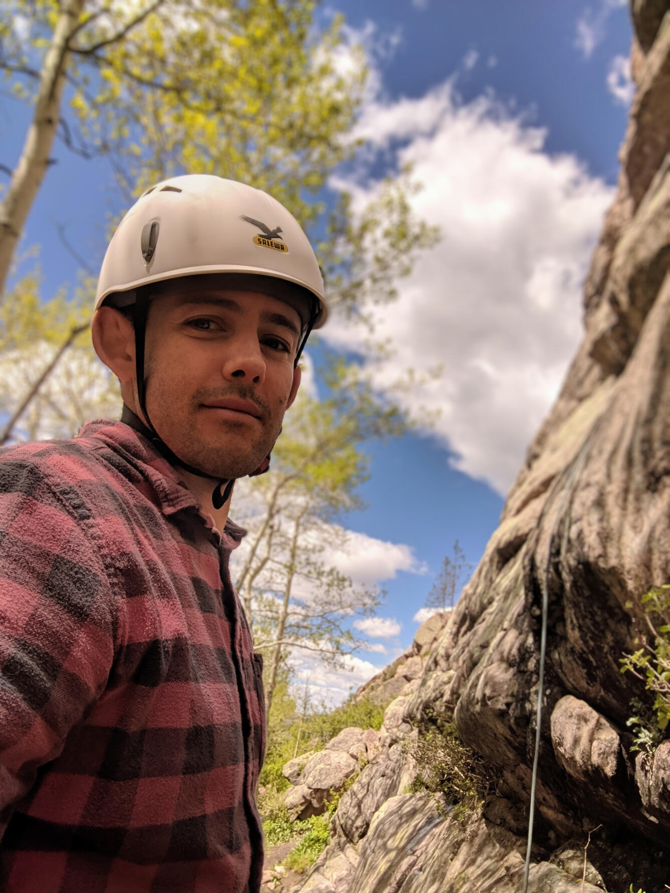 scientist wearing a white hat, red/black plaid shirt, near a rock outcropping and trees, on a sunny day