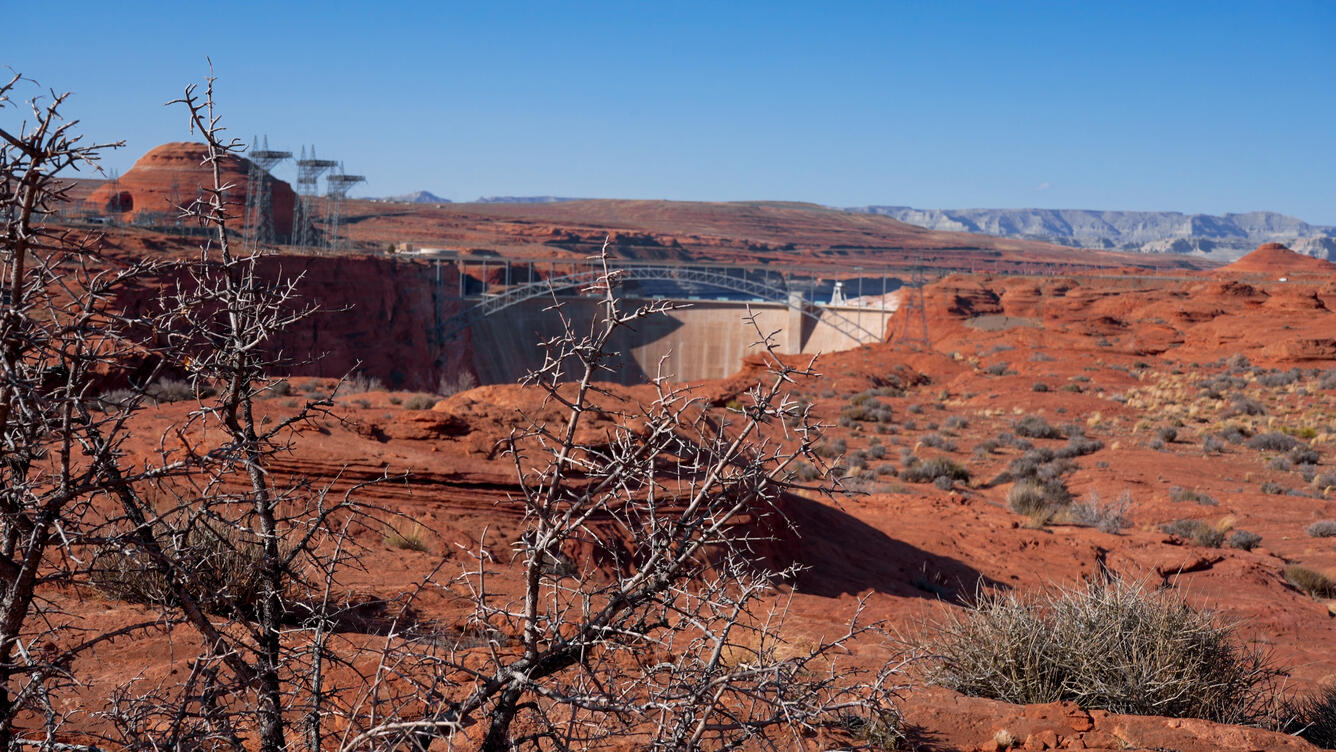 Red rocks and dry, sharp shrubs provide a backdrop for a concrete dam holding back hundreds of feet of water.