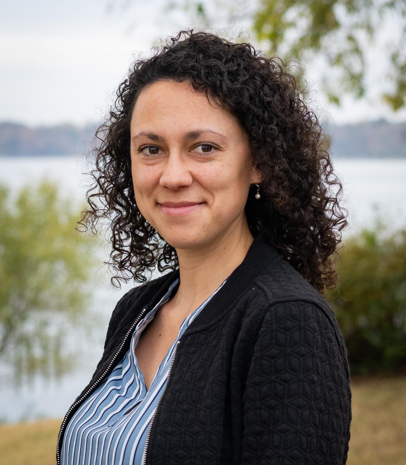 A woman with dark, curly hair in a blue shirt and black blazer stands for her picture.