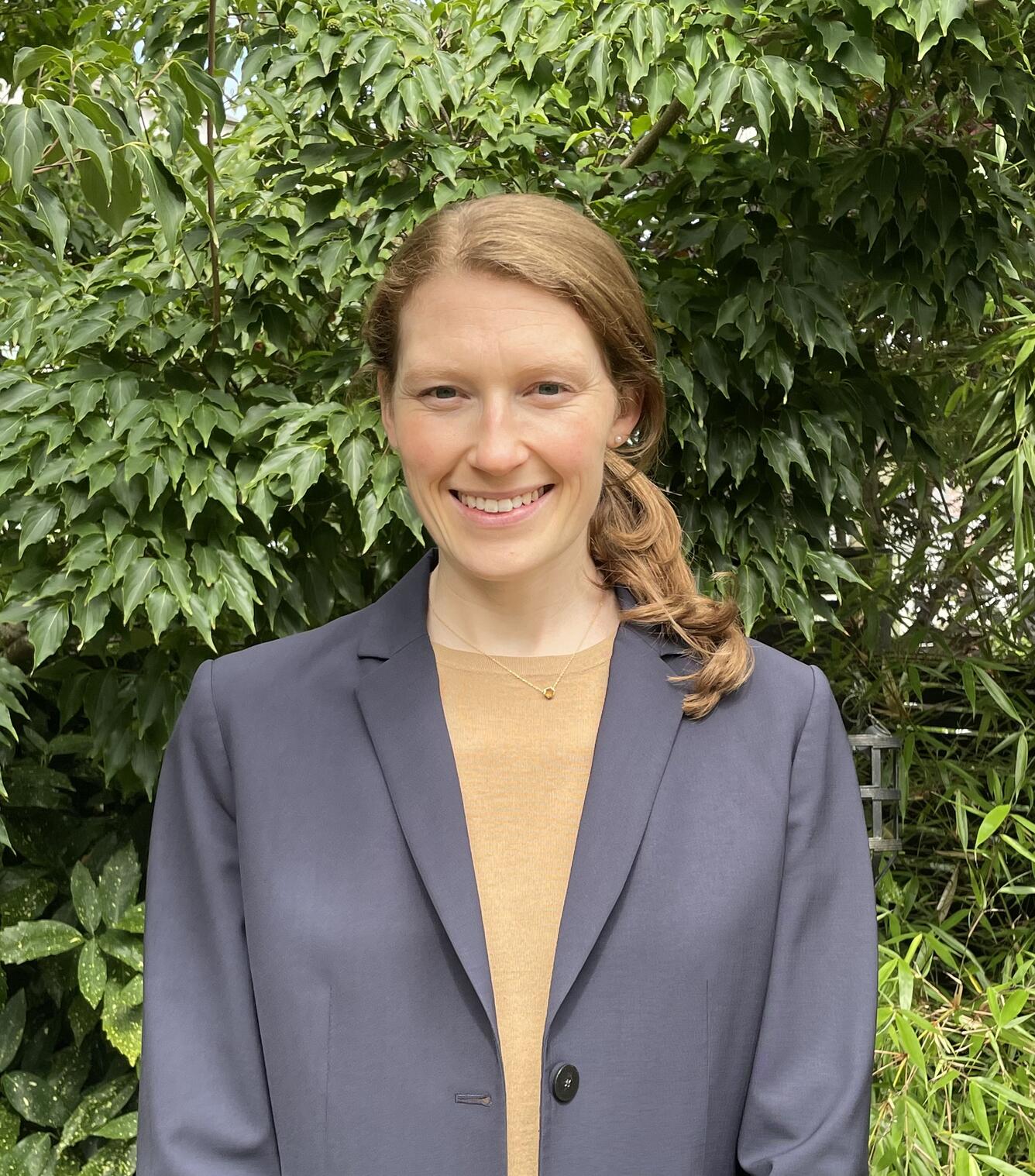 Woman in grey jacket and tan shirt standing in front of green plants.