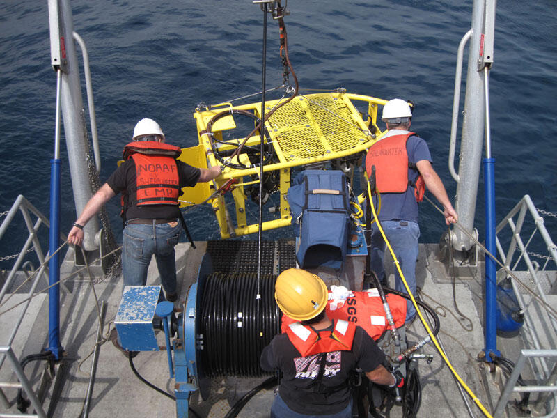 Three people guide a big metal frame, suspended with a cable and holding instruments, into the water off the deck of a boat.