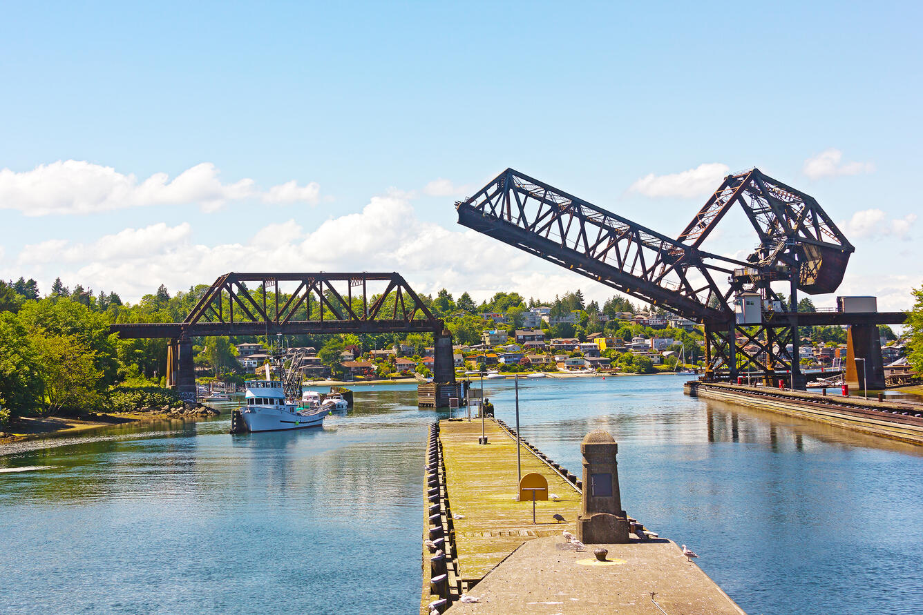 An red iron bridge crosses two cement lined canal channels. One side of the bridge is raised to allow passage. Partly cloudy