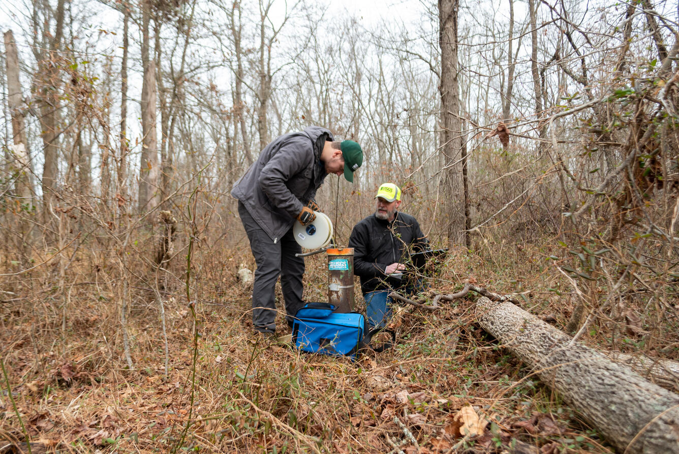 In a forest, one person measures a groundwater well while the other one inputs data into a laptop.