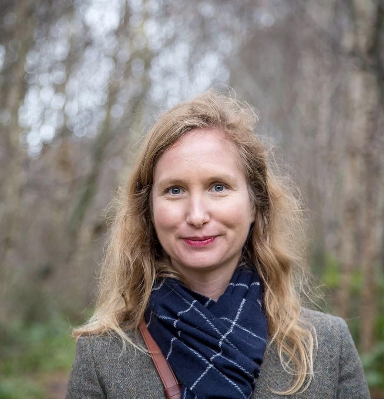 A woman with blonde hair wearing a jacket and scarf with trees in the background.