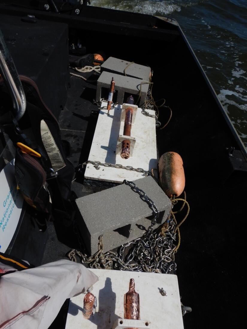 Two white rectangular platforms mounted with metal cylindrical objects sit on the deck of a boat chained to cinderblocks