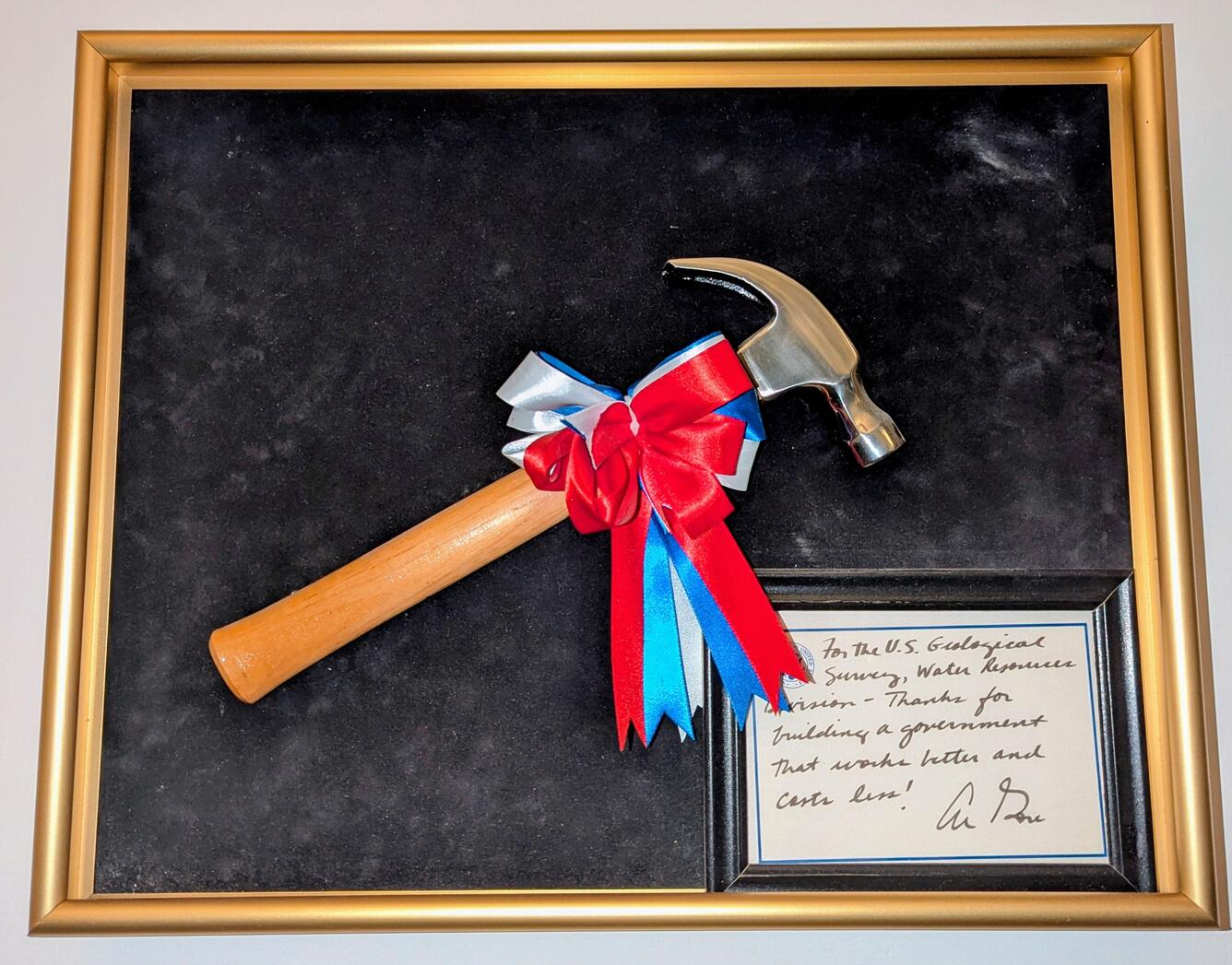 Hammer with a red, white, and blue bow mounted on black felt canvas with a golden frame