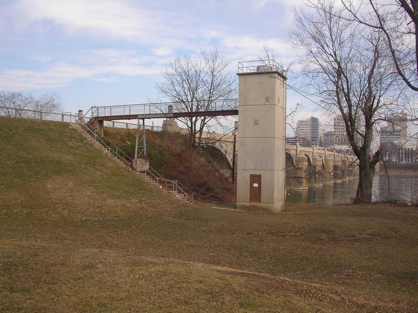 Photograph of narrow concrete tower with gangplank next to a river with a city in the background