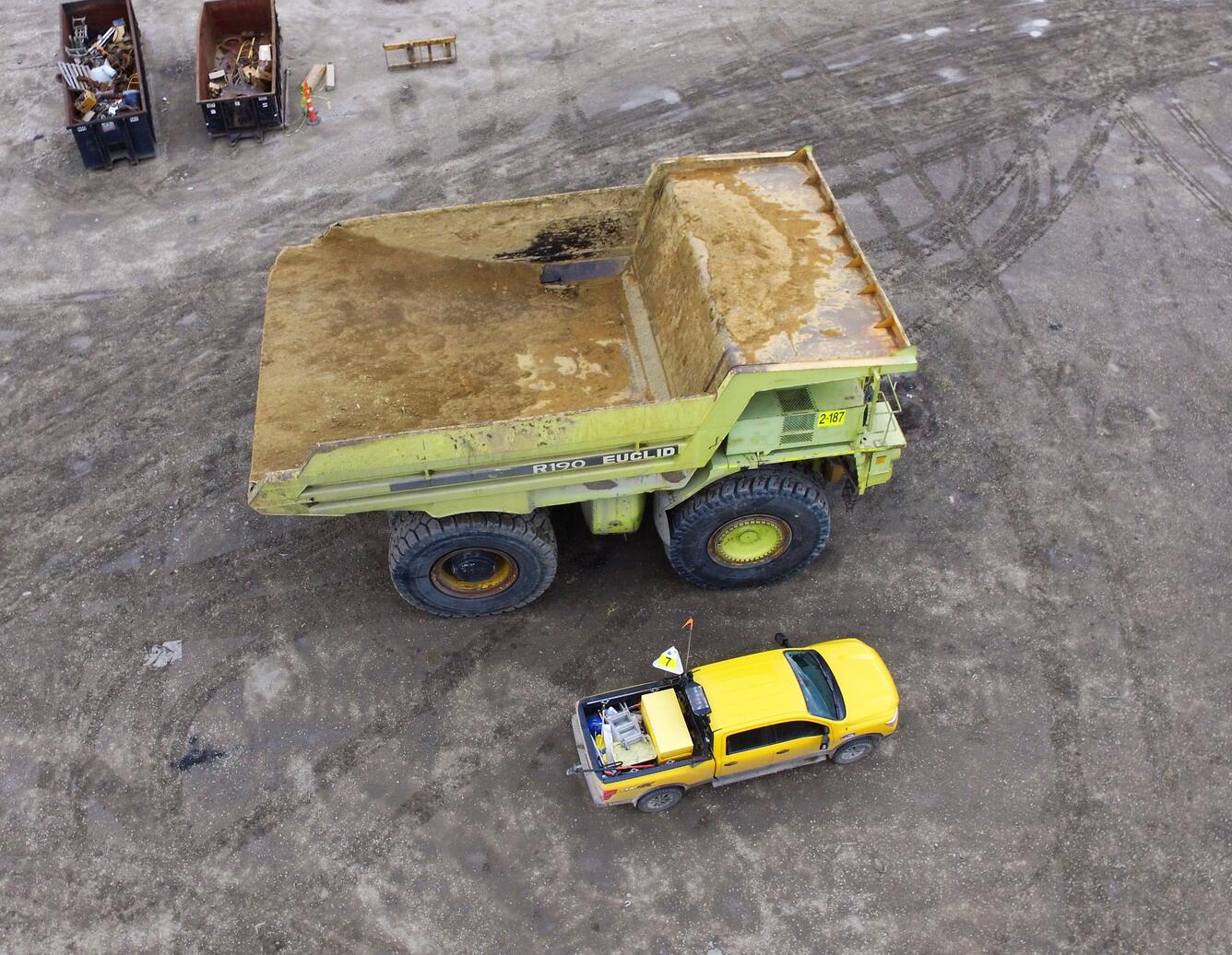 A drone image looking down on a very large truck parked next to a pickup that it dwarfs, with a couple of dumpsters
