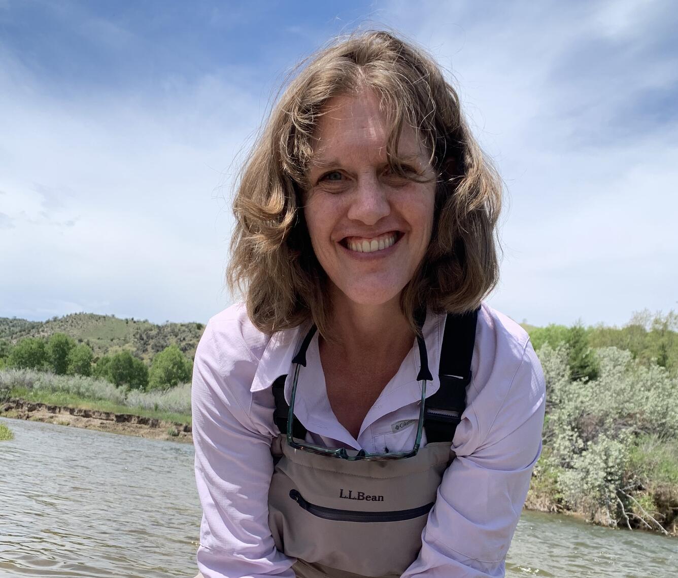 Anne smiling on a nice day against a backdrop of flowing river and plant covered river bank