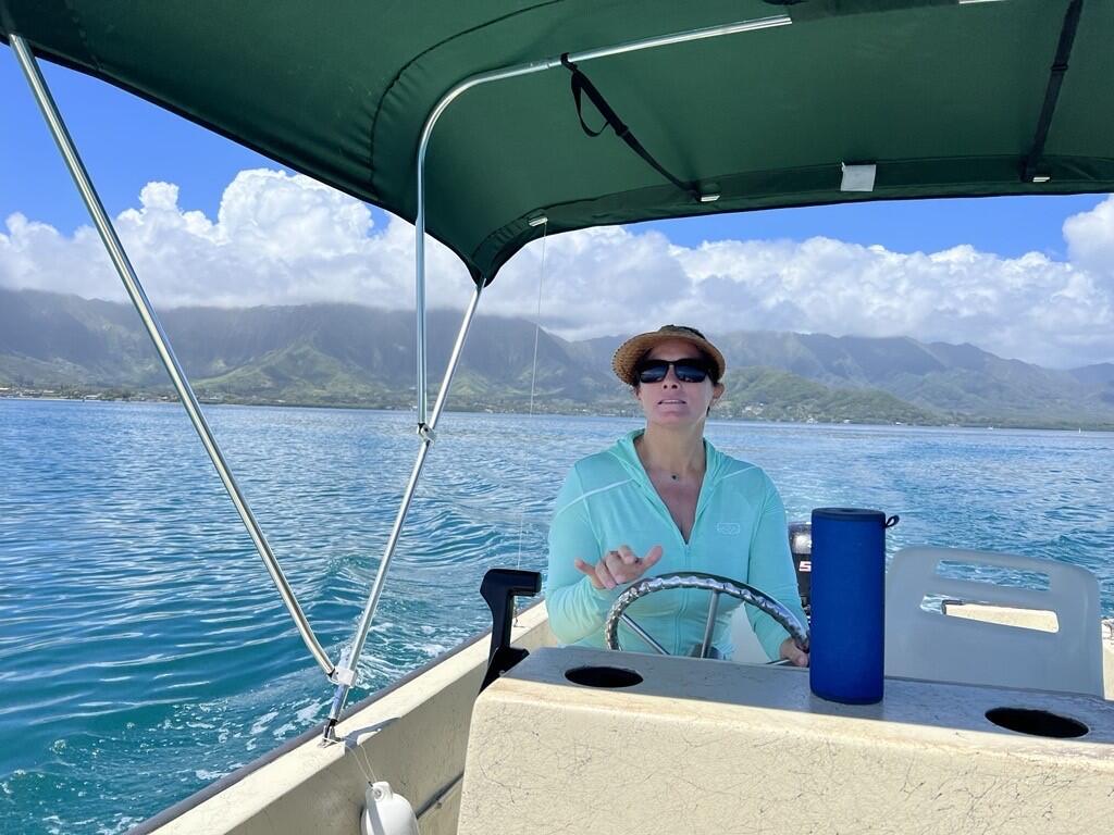 A woman wearing sunglasses and a hat drives a boat away from a lush Hawaiian island