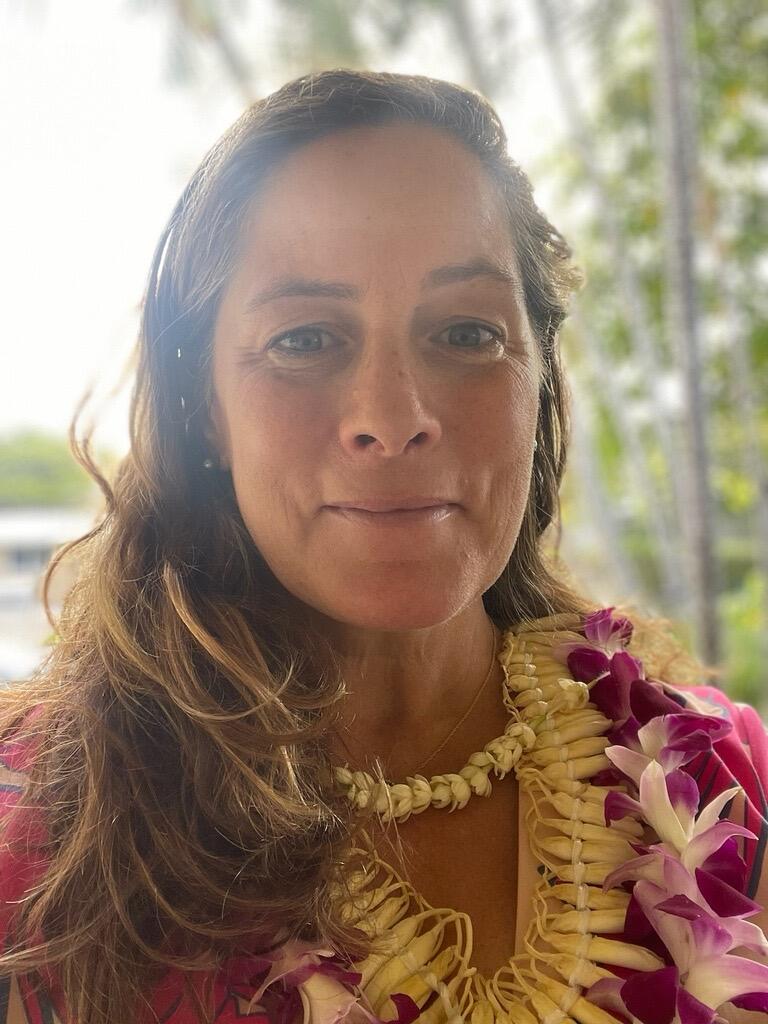 A woman with brown shoulder length hair wearing a flower lei and a bright floral dress