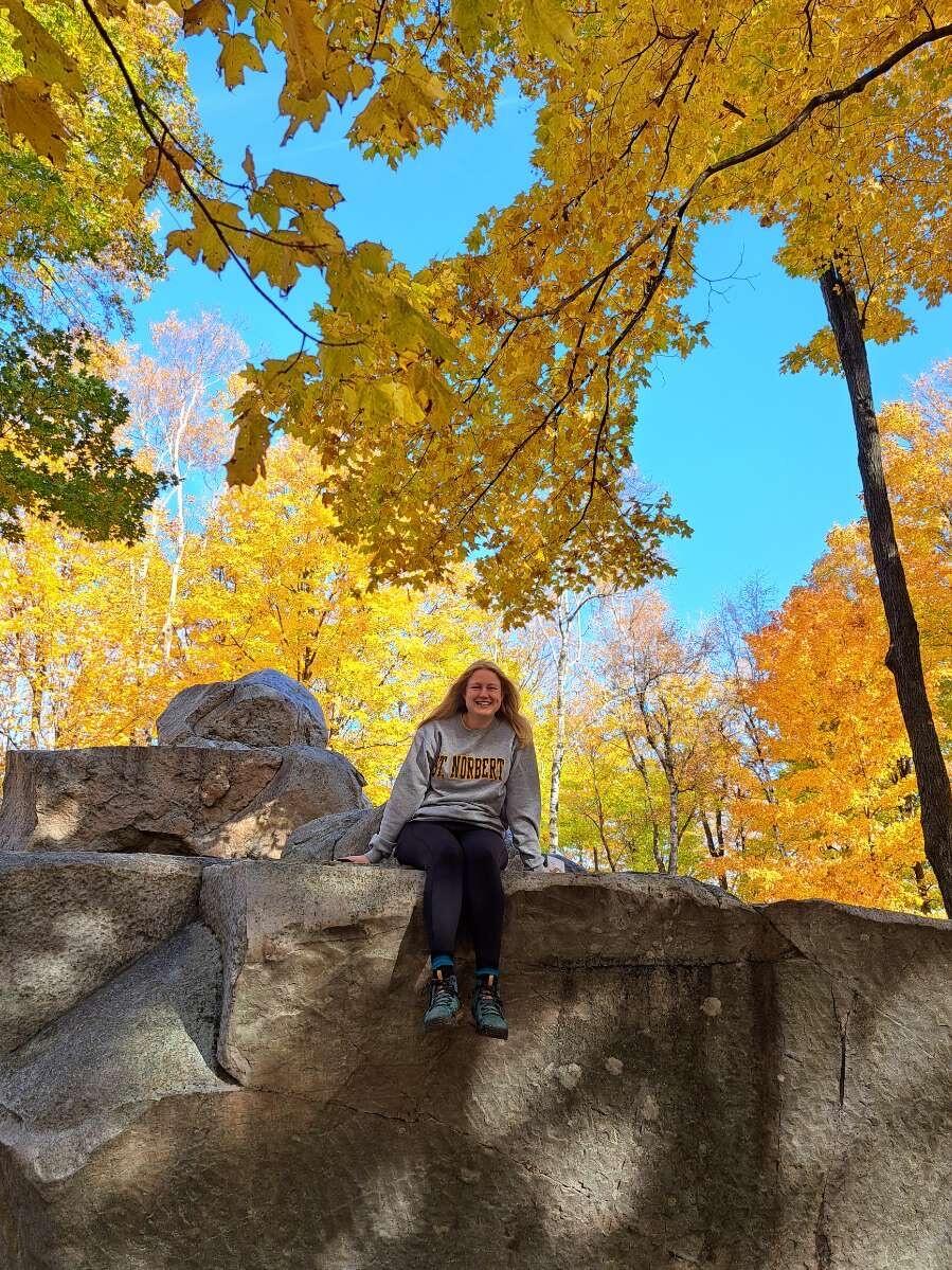 Woman sitting atop a rock formation with trees in fall colors in the background.