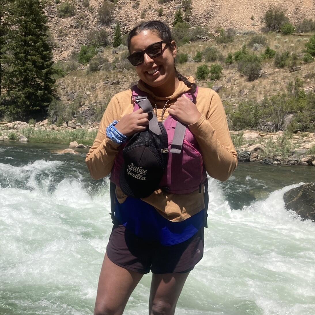 A smiling woman wearing a life jacket and sunglasses perches on a rock in front of river rapids