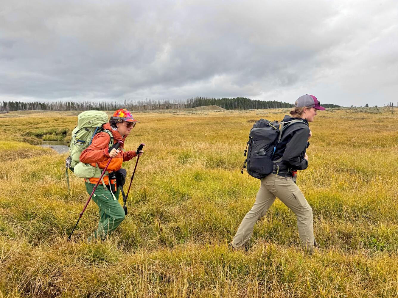 Two people wearing raincoats and hiking across a grassy meadow under cloudy skies