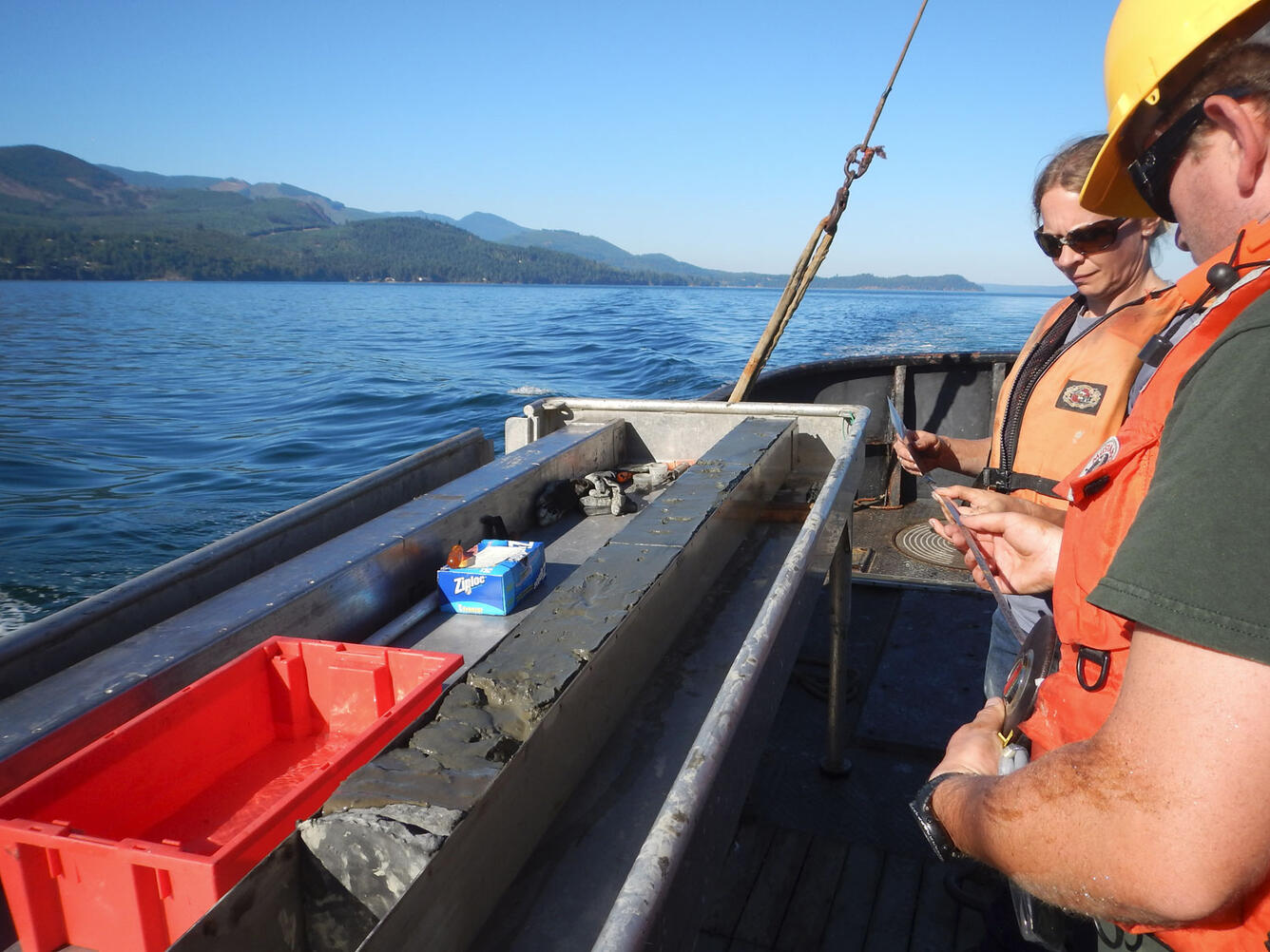 Two people hold a measuring tape standing near a long square core of mud on a boat.