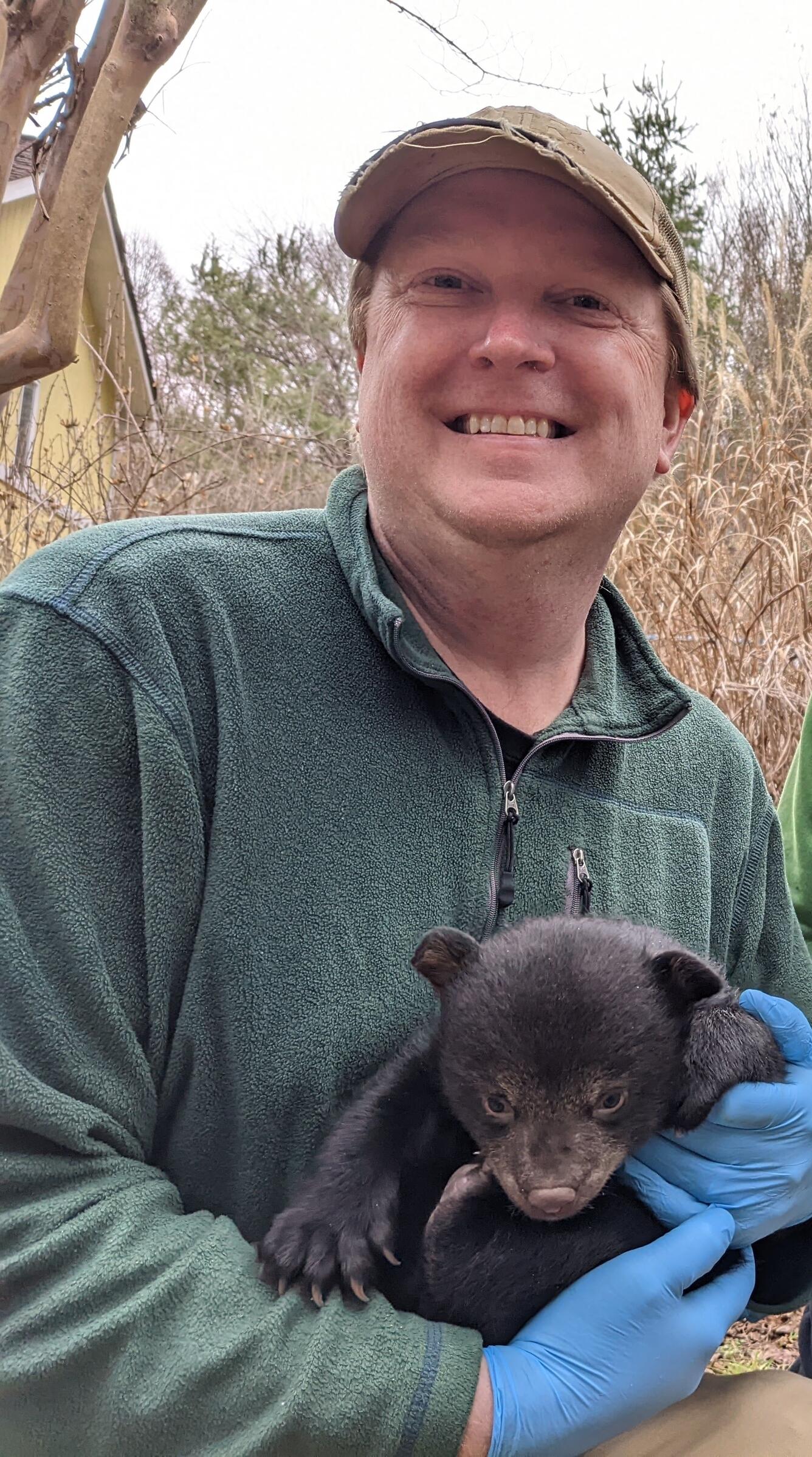 man standing in the woods, wearing a green sweatshirt and tan hat, holding a black bear cub