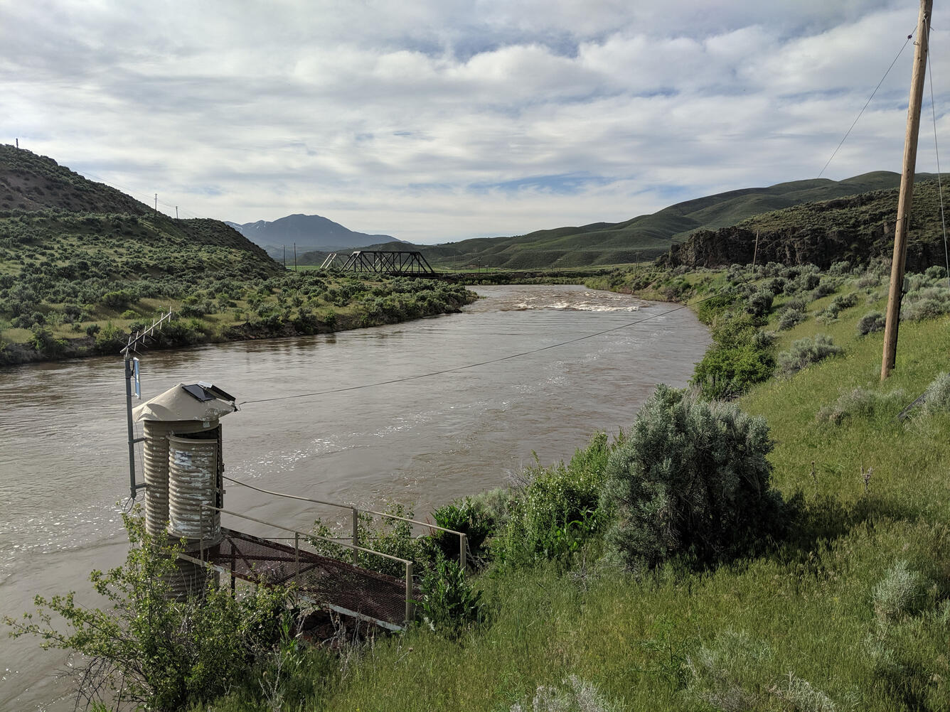 USGS gage house on the bank of the Humboldt River, with green hills surrounding the site and a cloudy sky overhead.