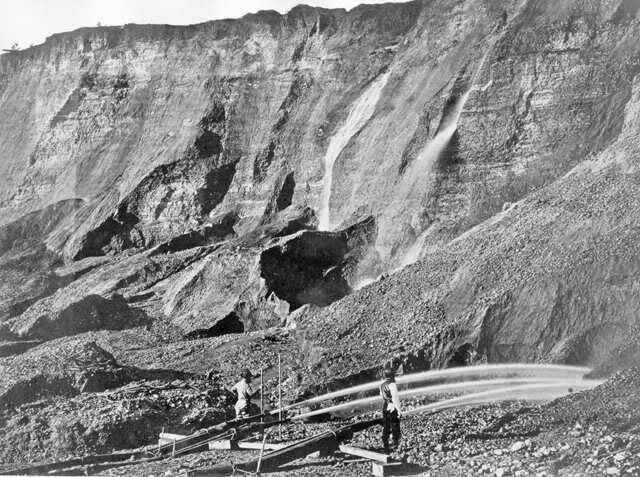 Vintage photo shows two men standing near jets of water that are aimed into a rocky slope.
