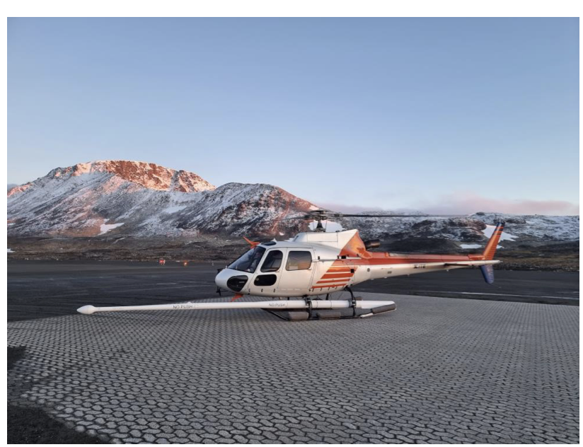 Red and white helicopter with boom extended, on surface in front of mountains.