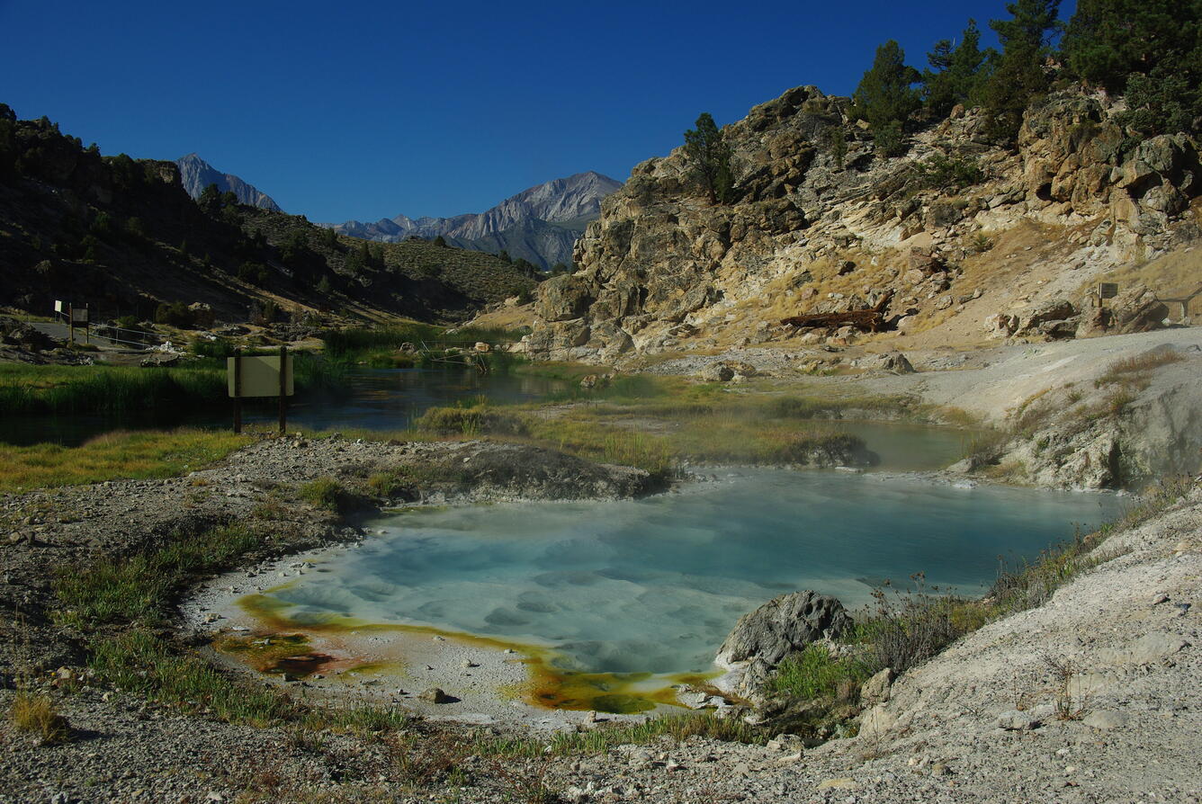 A sweeping view looks out over a clear blue hot spring and a creek bordered by bright green grass. In the background, rocky cliffs are scattered with sagebrush and pine trees. In the distance, bare, pointed mountains rise high into the air. The spring in the foreground is bordered by bleached white soil and steam rises from its surface.