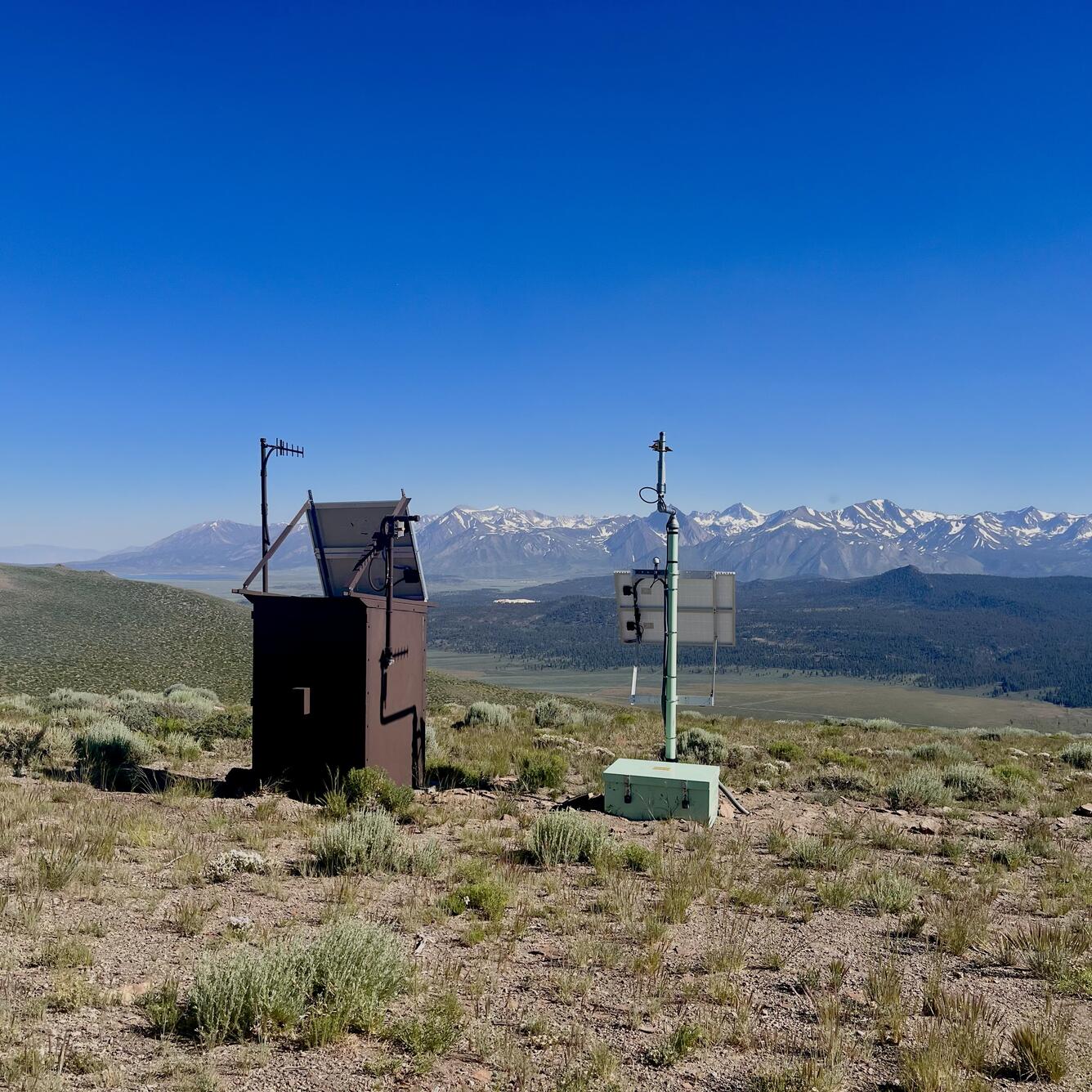 In this photo, a large brown metal box and a small aqua-colored box are both topped by antenna poles and solar panels. They sit side-by-side on a sagebrush-covered hill overlooking a broad valley and a line of jagged, snow-capped mountains.