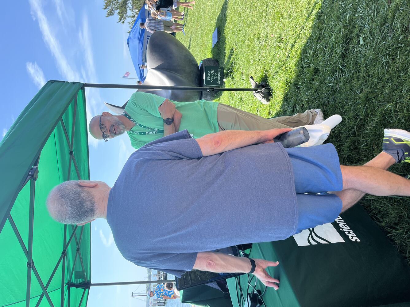 USGS scientist talking to a man next to a table, under a tent, outside by the water.