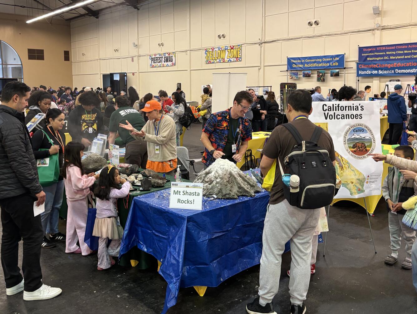This photo shows a science festival booth surrounded by children and their families. A model volcano sits on a blue tarp behind a "Mt Shasta Rocks!" sign. Behind the table, a scientist in an orange USGS cap explains volcanic rocks, while another scientist in a volcano-themed Hawaiian shirt prepares the model volcano for its next "eruption".