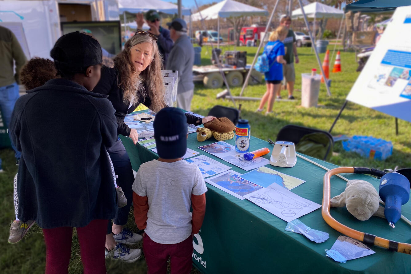 USGS staff points to a toy turtle on a table outside while speaking to a family at an outreach event