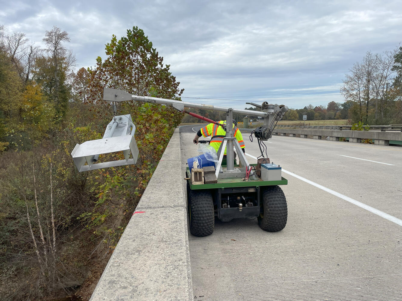 A bedload sampler suspended by a reel over a bridge above a river. The sampler's reel is mounted on a utility vehicle.