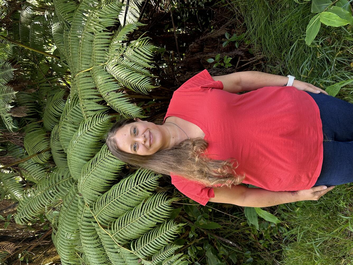 Person in orange shirt with long brown hair in front of tree ferns.