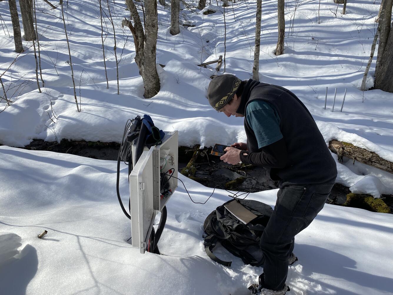 A man in the woods in the snow looks at a data logger.