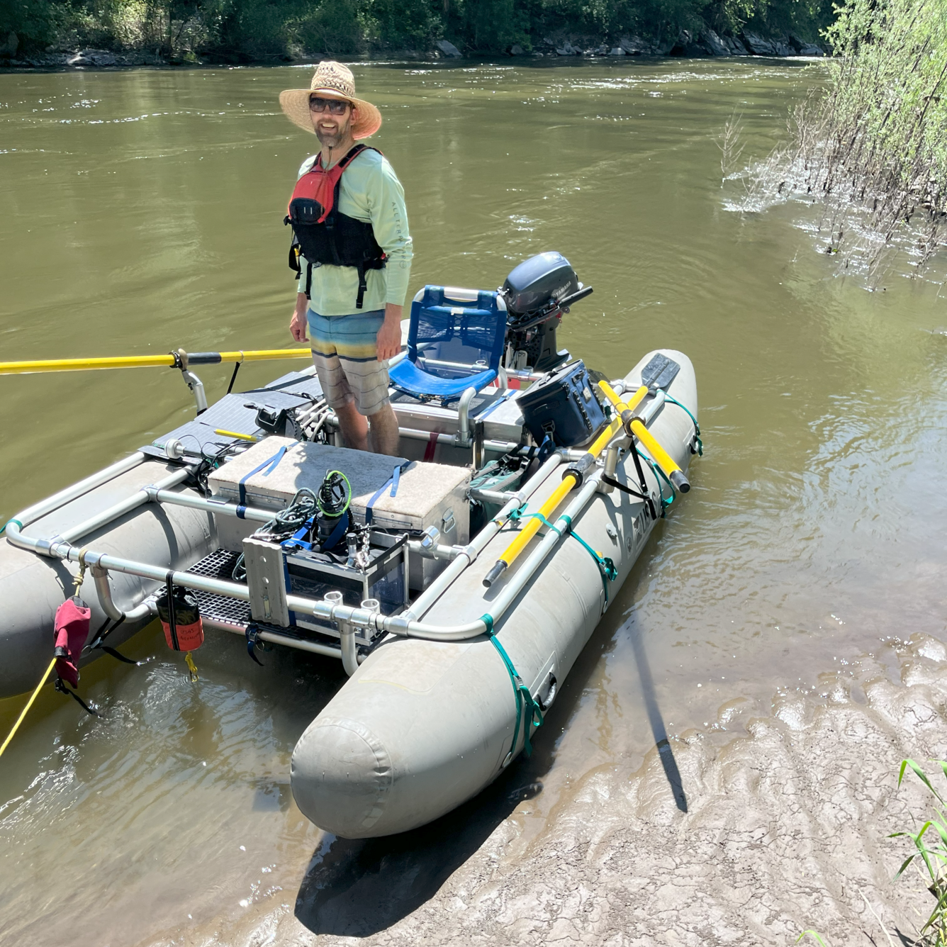 Man standing in an inflatable catamaran boat laden with equipment on the river bank 