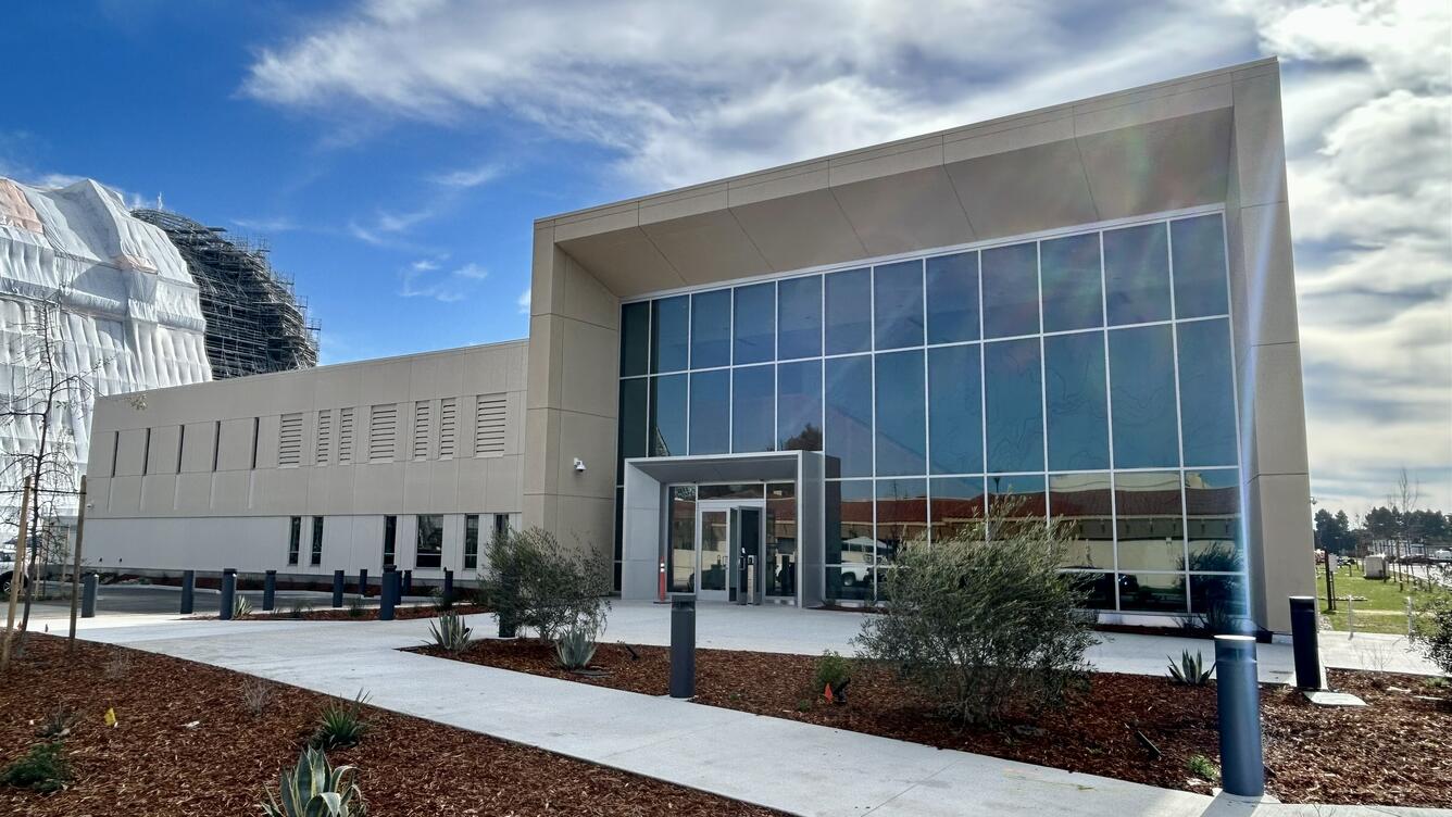 A rectangular beige building with a large, glass-fronted lobby is viewed at an angle. The building is surrounded by mulched landscaping and in the left background is a huge dirigible hangar under construction.