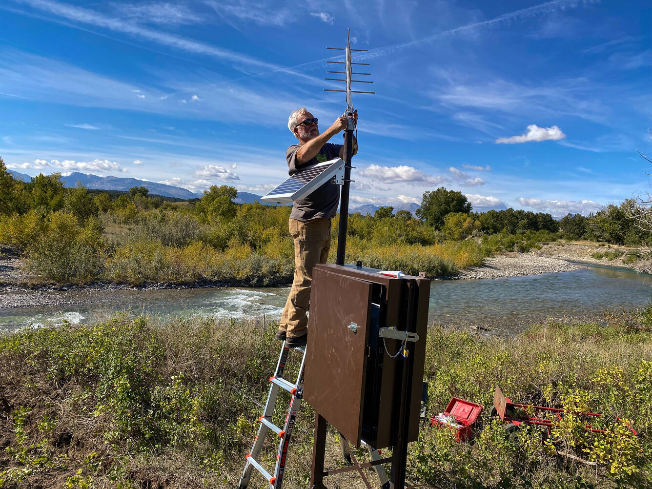 Hydrologist on a ladder installing wire to antenna connected to streamgage box with creek in background 