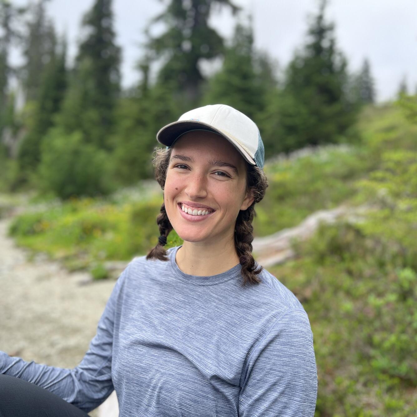 Callie is in a grey long sleeved shirt with a white ball cap and brown hair braided on either side of her face.