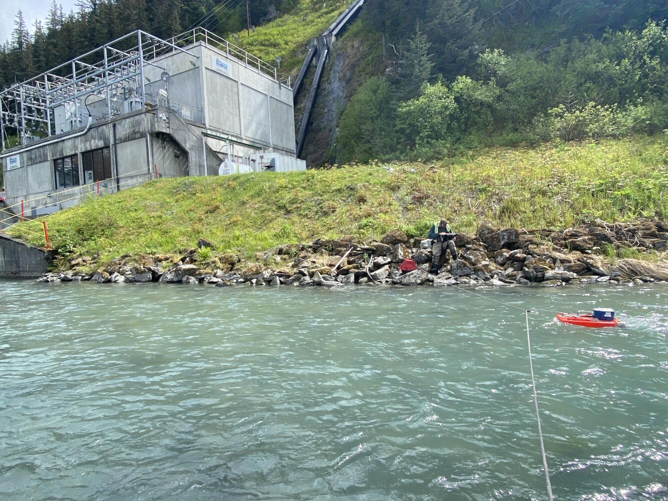 Scientist sitting on rocks. Tethered Acoustic Doppler Current Profiler on water on right side measuring streamflow.