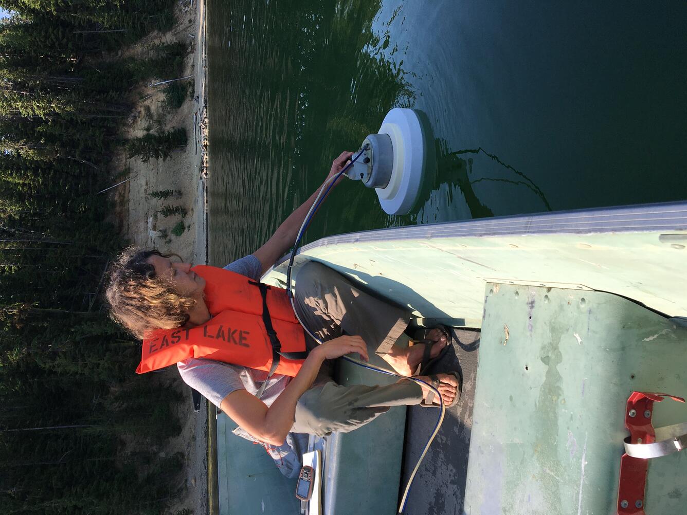 A woman in a life jacket sits in a small motorboat, holding a piece of gas sampling equipment in the water of a lake. The lake is surrounded by steep walls covered in tall pine trees.