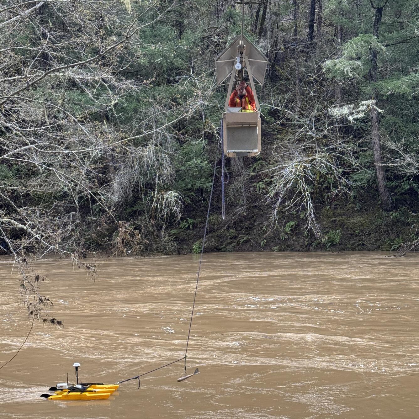 man pulls cable car across rushing brown water with a yellow pontoon in tow that has sensors recording the flow